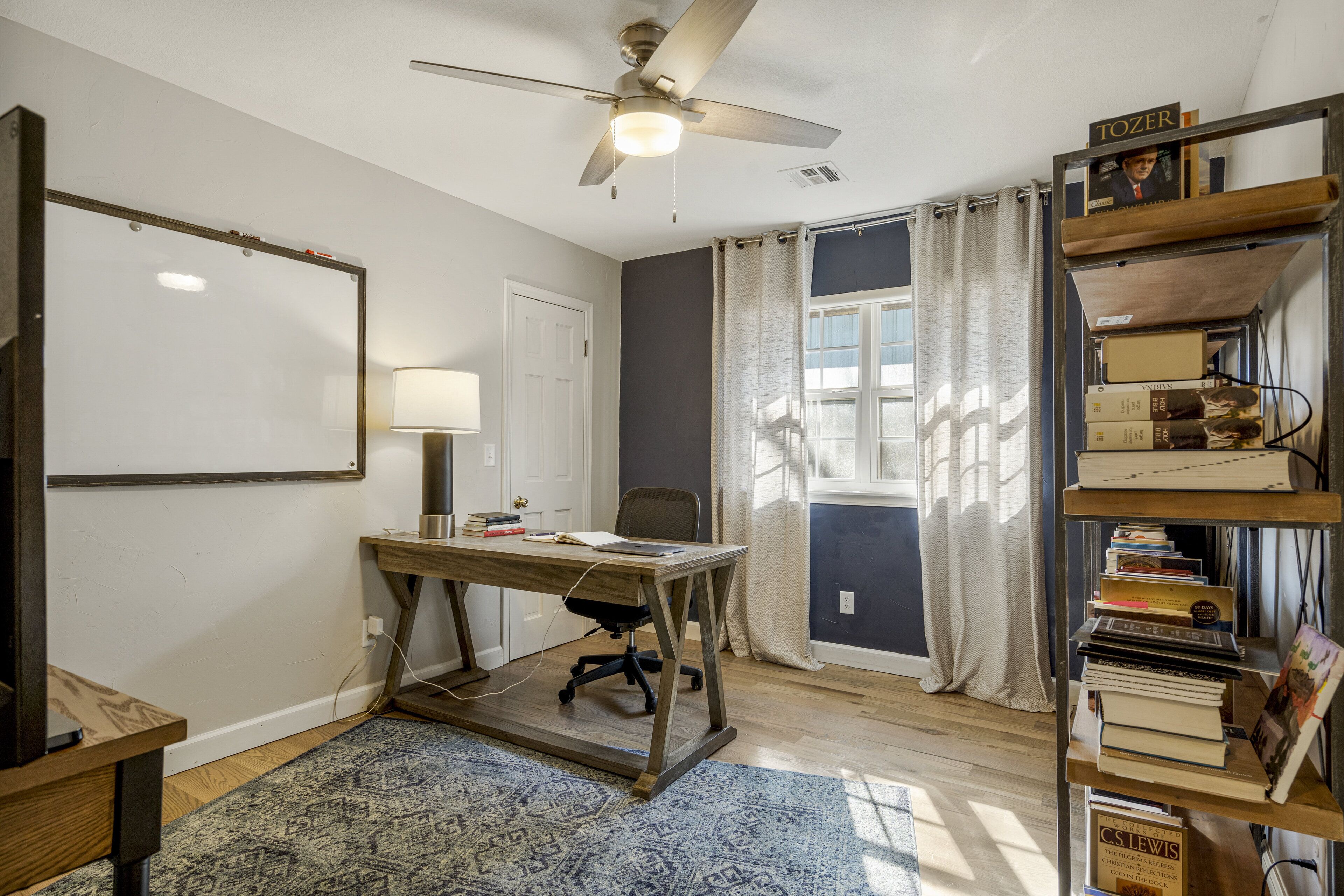 A room with a desk, chair, bookshelf, ceiling fan, and a window with curtains.