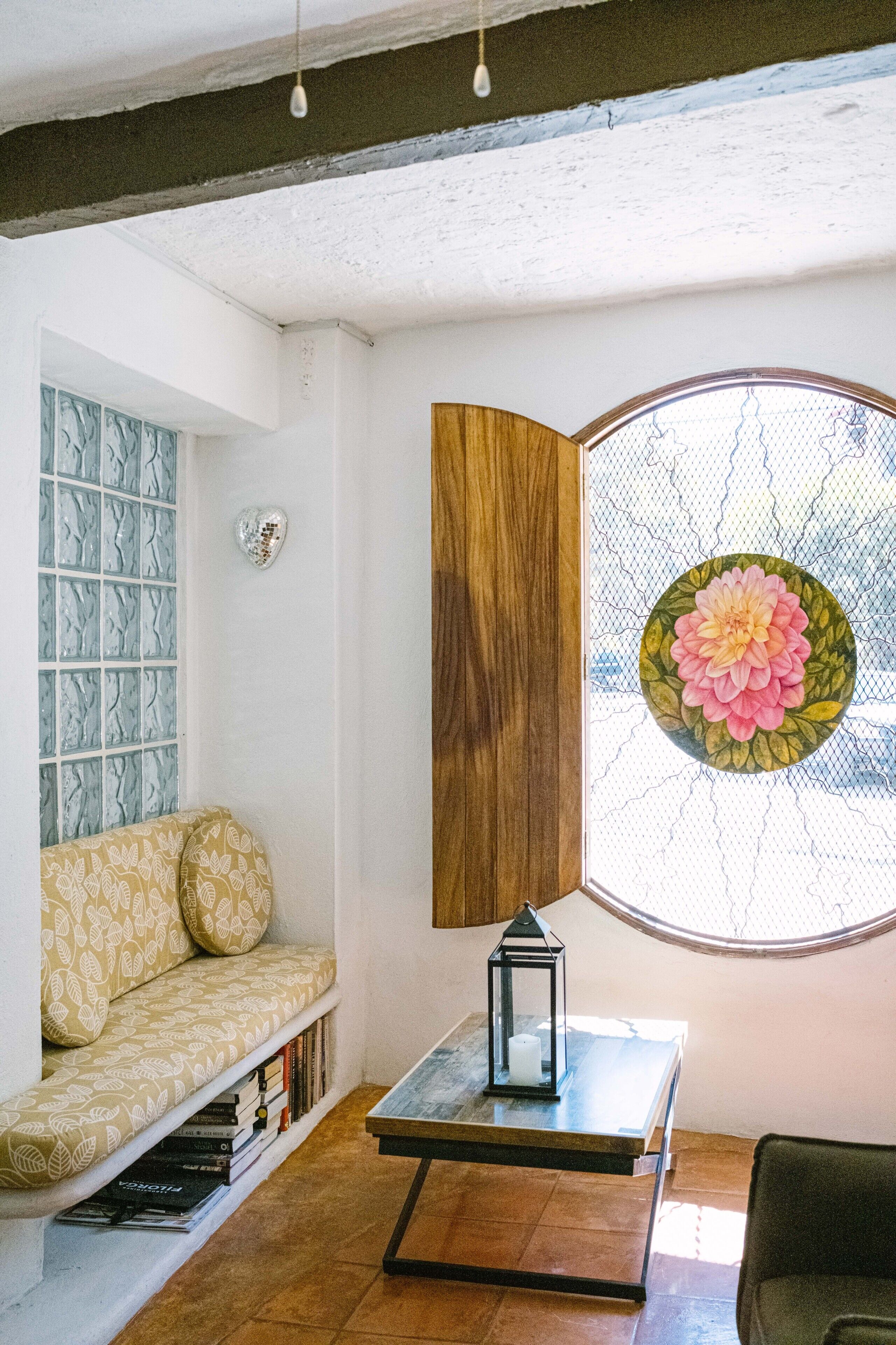 A cozy reading nook with a cushioned bench, a small table with a lantern, and a large window with a floral design.