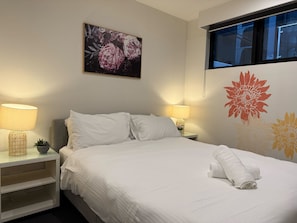 A neatly made bed with white linens, two bedside lamps, a small potted plant, and a floral painting on the wall.