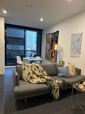 A modern living room with a grey sofa, a coffee table, and a large window with a city view. - City-View Chic on Bourke | Free Tram + Netflix (Melbourne)