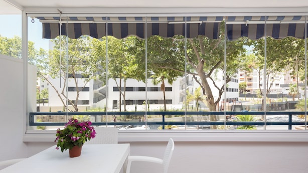 Un balcón con una mesa y sillas blancas, una planta en maceta y vistas a edificios y árboles.