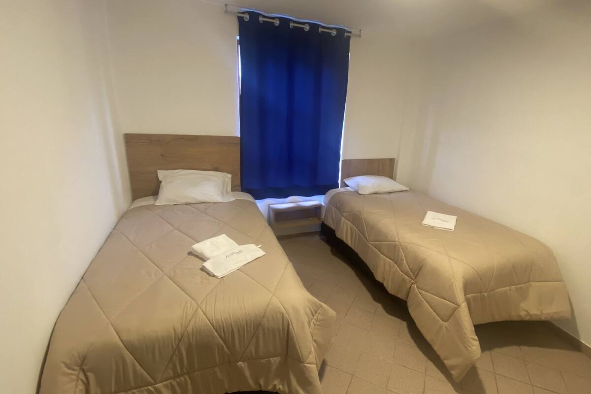 A small, clean room with two single beds, a wooden headboard, a window with blue curtains, and a small table between the beds.
