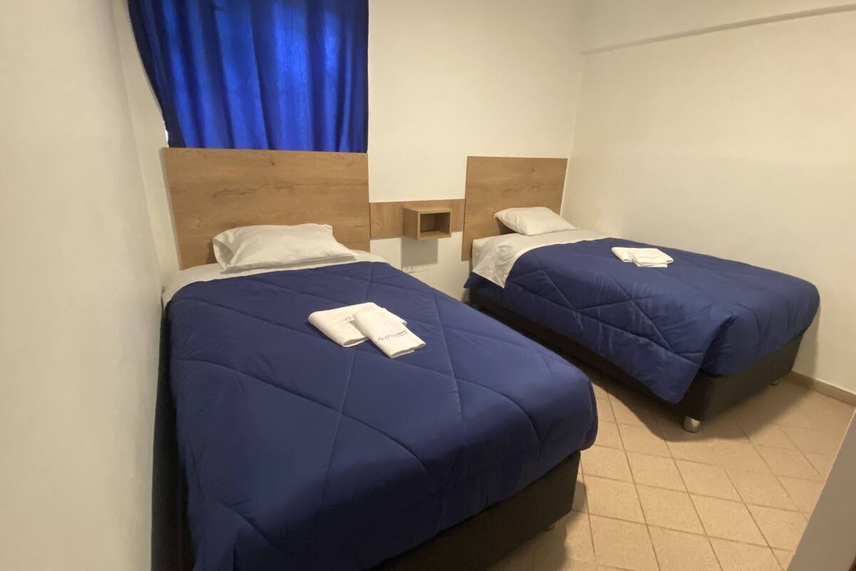 A room with two beds, each with a blue bedspread and white pillows. There is a wooden headboard, a small bedside table with a lamp, and a blue curtain.