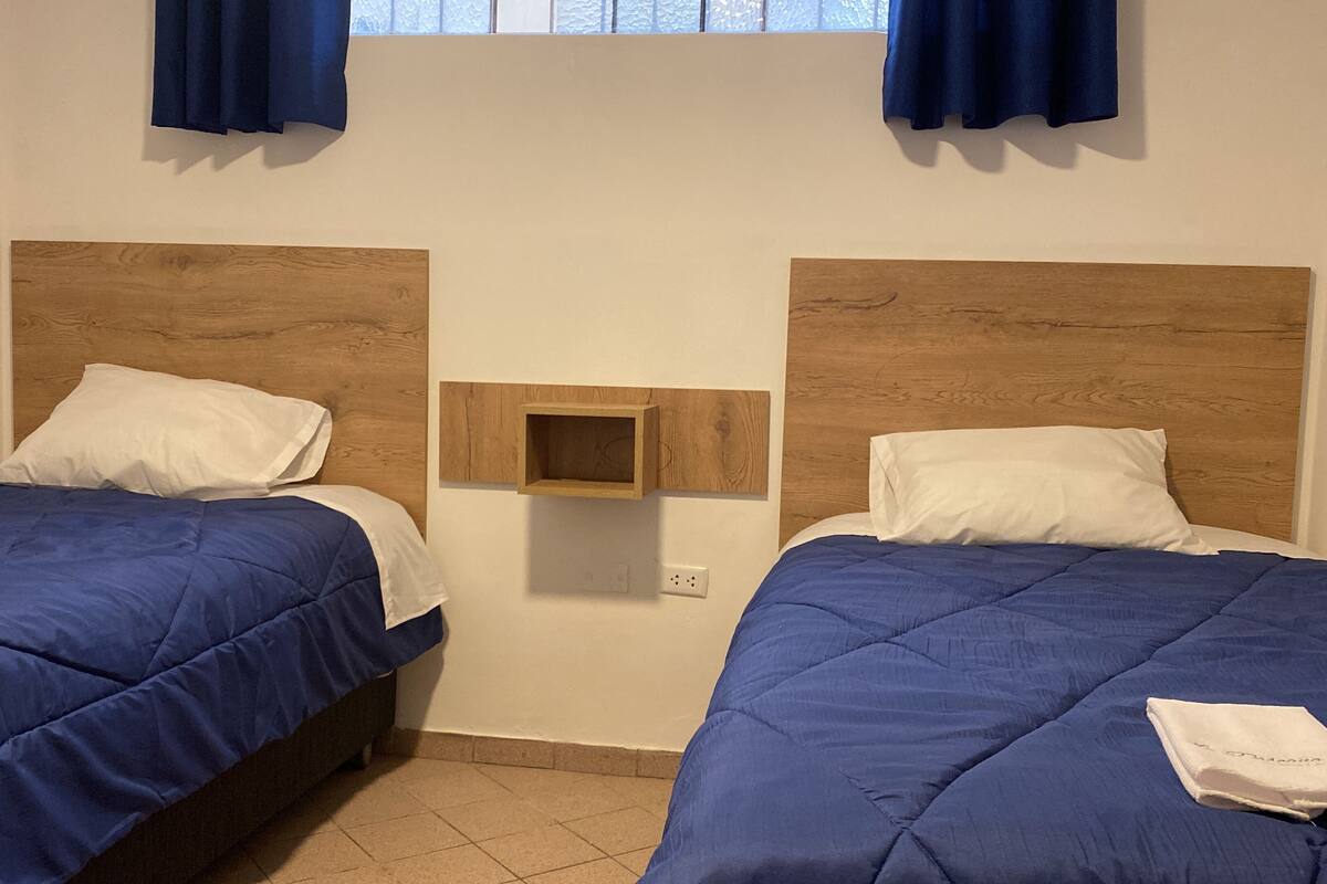 A double room with two beds, each with a wooden headboard, a small wooden shelf, and a window with curtains.