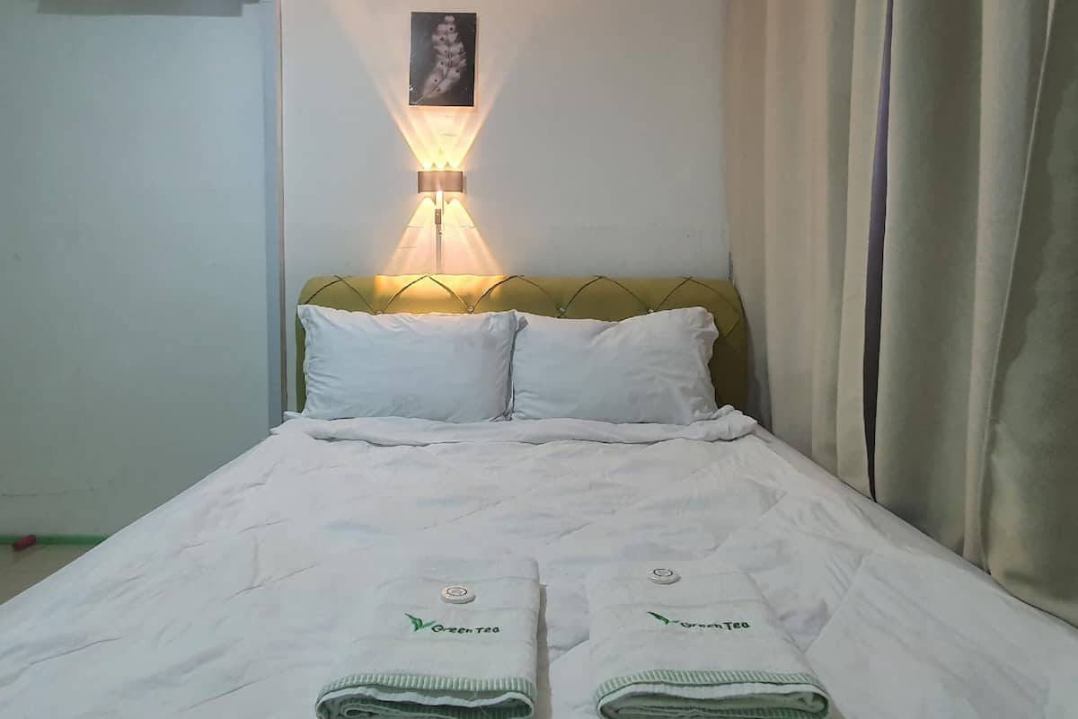 A hotel room with a bed, a wall-mounted air conditioner, a wall-mounted lamp, and a framed picture on the wall.