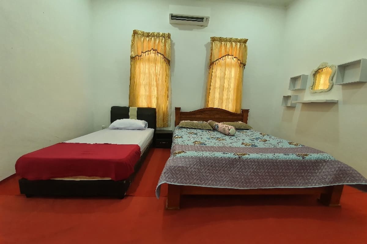 A room with two beds, one with a red bedspread and the other with a patterned bedspread. There is a wall-mounted air conditioner above the beds, and a shelf with decorative items on the wall.