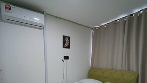 A single-bed room with a yellow headboard, white bedding, a wall-mounted air conditioner, a small framed picture, and a curtain on the window.