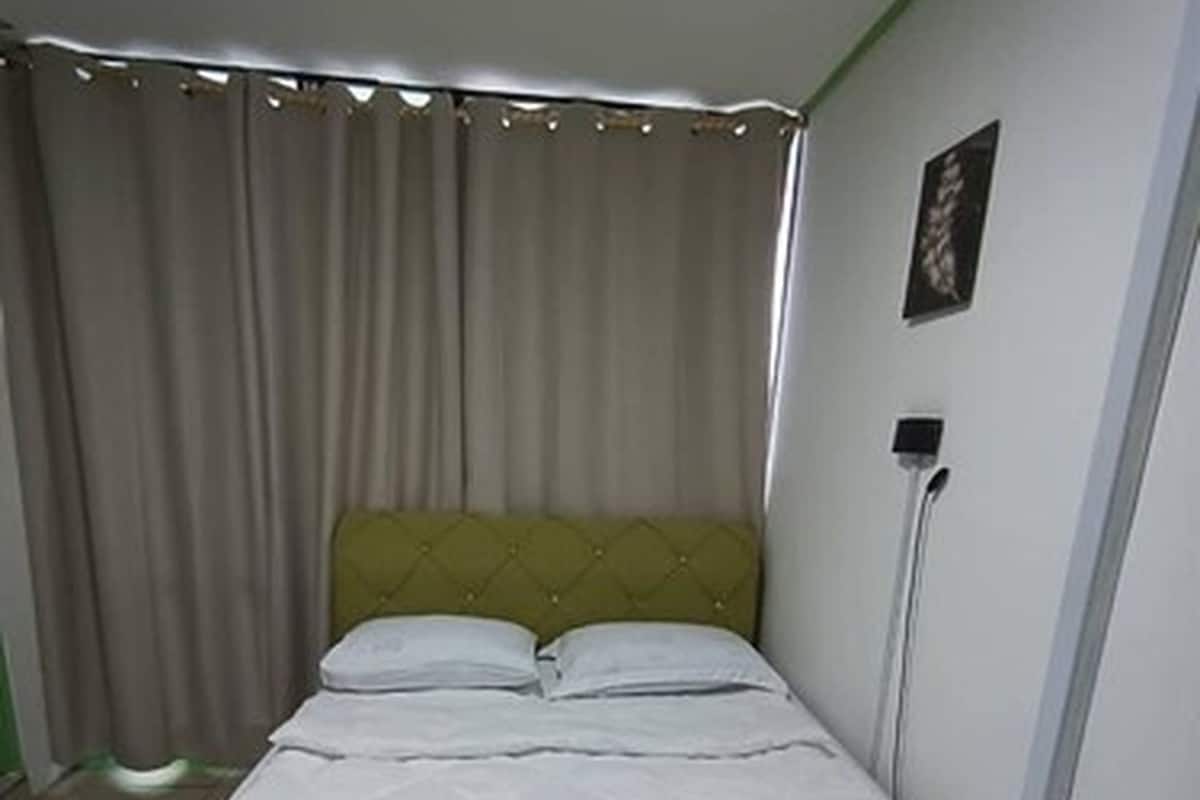 A single bed with a quilted white bedspread, a green headboard, and a white wall with a power outlet.