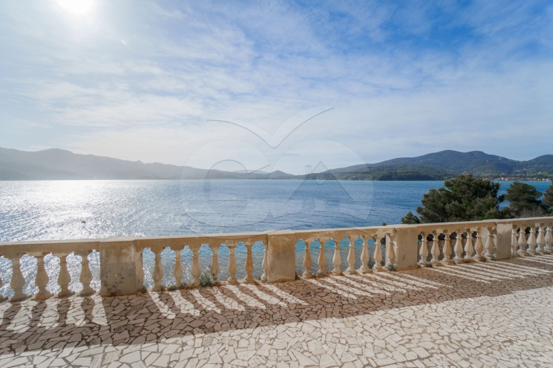 A coastal view with a tiled walkway and balustrade overlooking the sea and distant mountains.