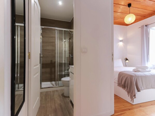 A modern bedroom with a wooden floor, a glass shower enclosure, a white toilet, and a neatly made bed with a lamp and towels.