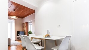 A modern dining area with a white table and four white chairs, a wooden ceiling, and a view into another room with a television.
