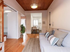 A modern living room with a wooden ceiling, a light-colored sofa with striped cushions, a large plant, and a dining area in the background.