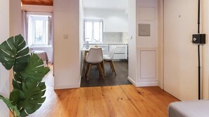 A modern interior with a wooden floor, a dining area with a white table and chairs, and a kitchen visible through an open door.