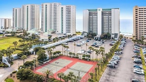 A multi-story hotel with a parking lot, tennis courts, and a view of the ocean.