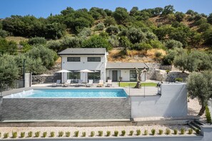 A modern two-story house with a swimming pool, outdoor seating, and a view of a hillside.