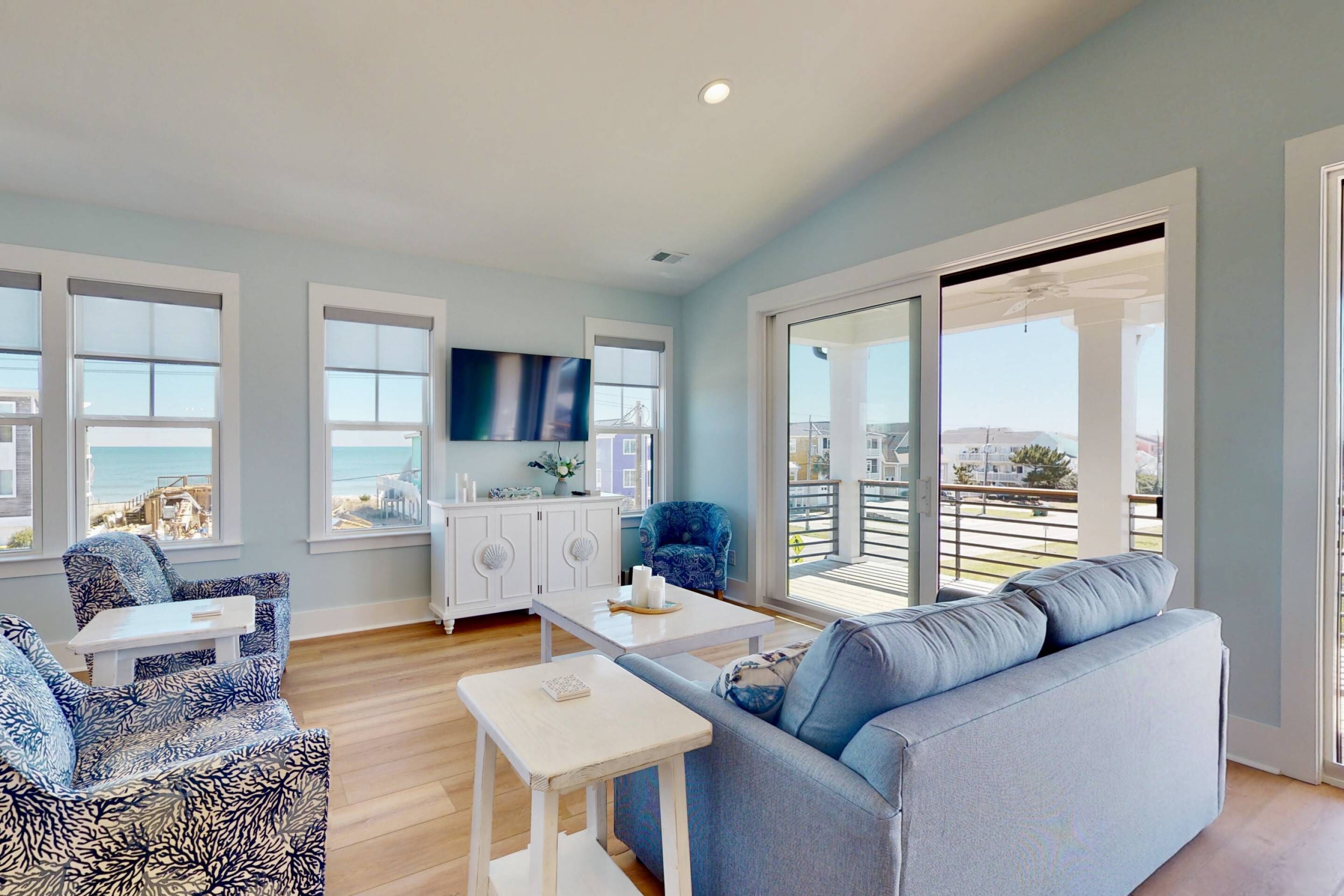 A spacious living room with a blue sofa, patterned armchair, and a white coffee table. A flat-screen TV is mounted on the wall, and there is a large sliding glass door leading to an outdoor balcony.