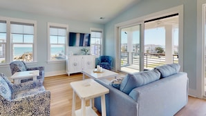 A spacious living room with a blue sofa, patterned armchair, and a white coffee table. A flat-screen TV is mounted on the wall, and there is a large sliding glass door leading to an outdoor balcony. - Luxury Kure Beach Getaway | Steps to Beach, Ocean Views, Elevator, Grill + Games (Kure Beach)