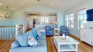 A spacious living room with a blue sofa, patterned armchairs, and a white coffee table. A ceiling fan and pendant light are visible. - Luxury Kure Beach Getaway | Steps to Beach, Ocean Views, Elevator, Grill + Games (Kure Beach)