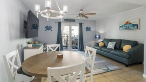 Un salon comprenant une table à manger, des chaises blanches, un canapé vert, un ventilateur de plafond et un panneau indiquant la direction de la plage.