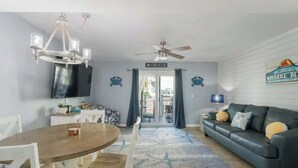 Un salon avec un canapé vert foncé, une table à manger en bois avec des chaises, un lustre, un ventilateur de plafond et un téléviseur fixé au mur.