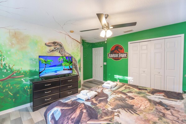 A bedroom with a green wall, a mural of a dinosaur, a bed with a dinosaur-themed bedspread, a television on a dresser, and a ceiling fan. - Awesome Movie Themed 6 Bedroom Villa, Private Pool/Spa in Gated Resort (Kissimmee)