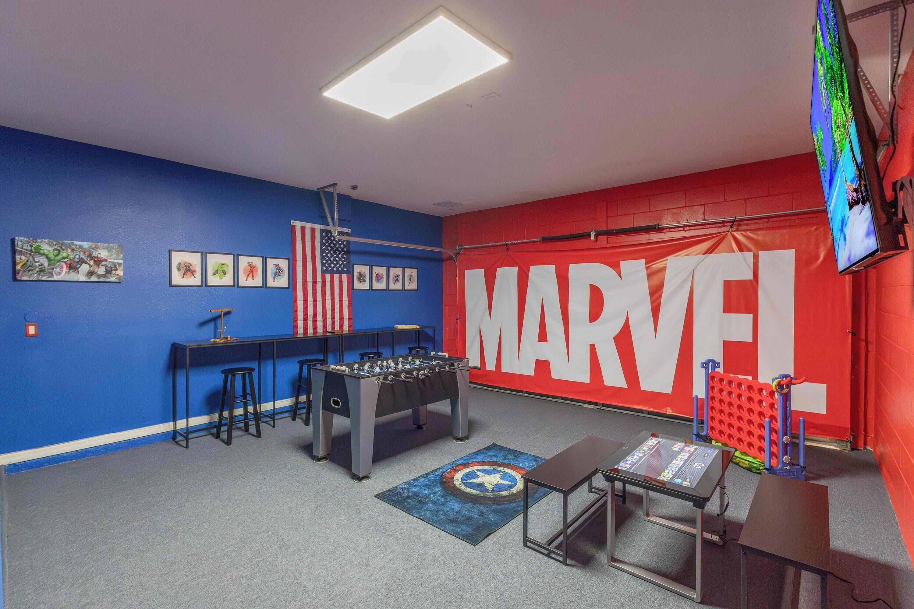 A room with a foosball table, a foosball table with a Star-Spangled Banner rug, and a large red wall with the word "MARVEL" on it.