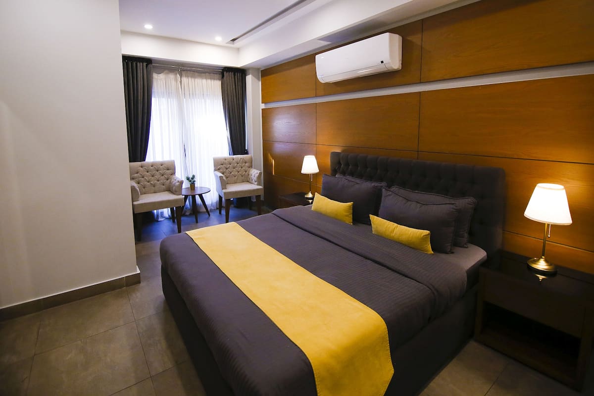A hotel room with a large bed, two bedside lamps, a small table with a chair, and a window with curtains.