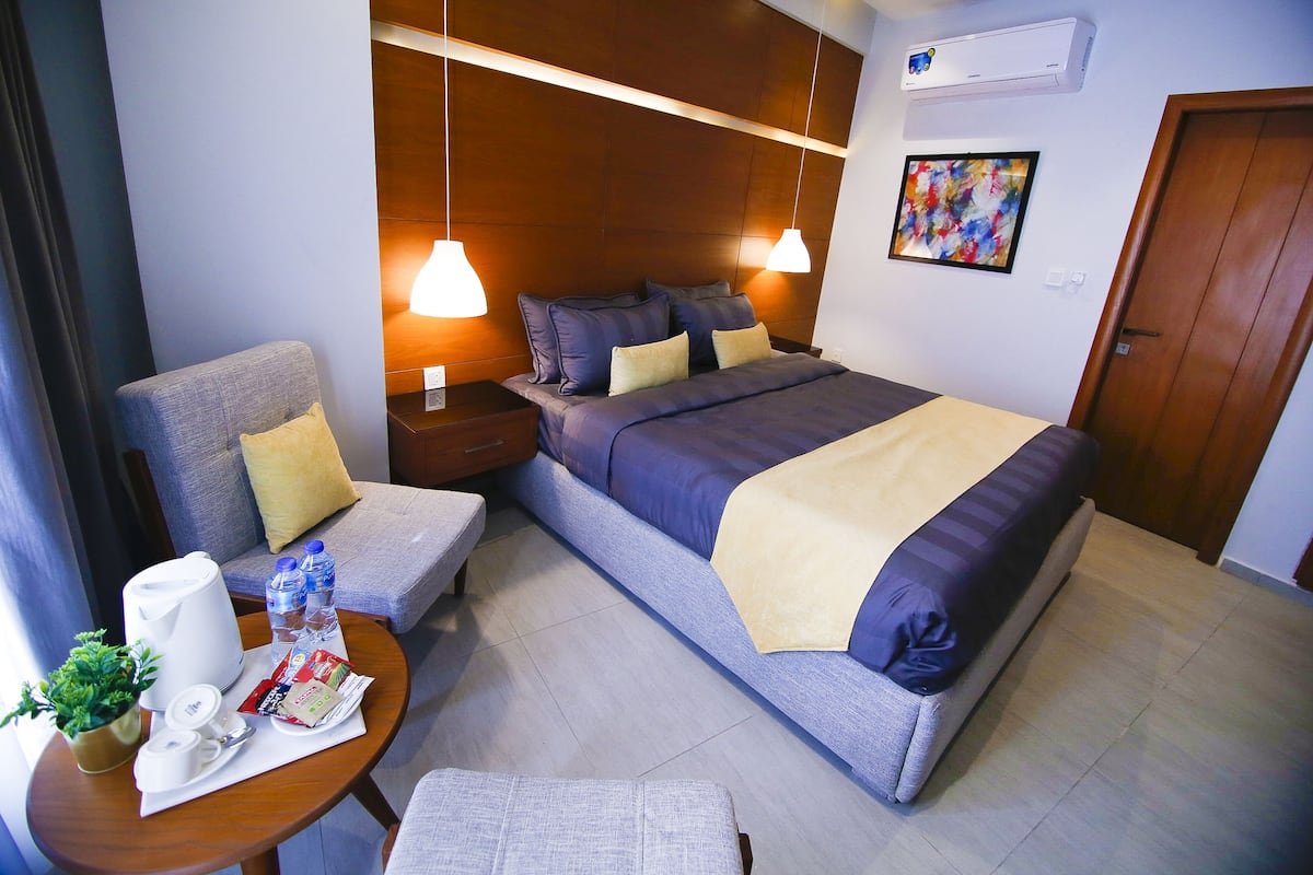 A modern hotel room with a large bed, a sofa, a small table with amenities, and a wooden headboard.