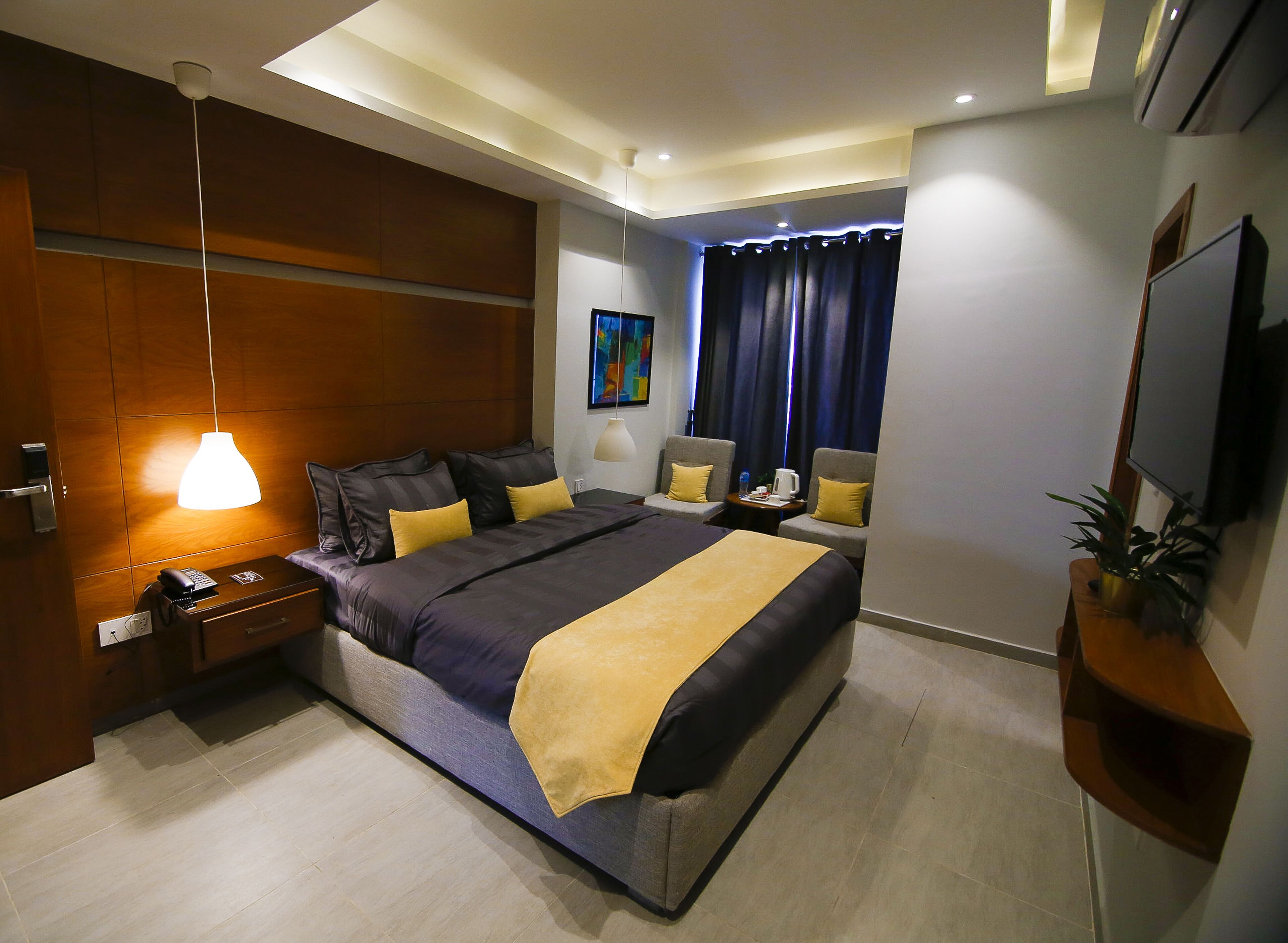A modern hotel room with a large bed, bedside tables, a desk, a chair, a desk lamp, a painting, a potted plant, and a flat-screen TV.