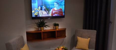 A living room with two grey armchairs, a small wooden table with a fruit bowl, a mounted TV, and a shelf with plants.