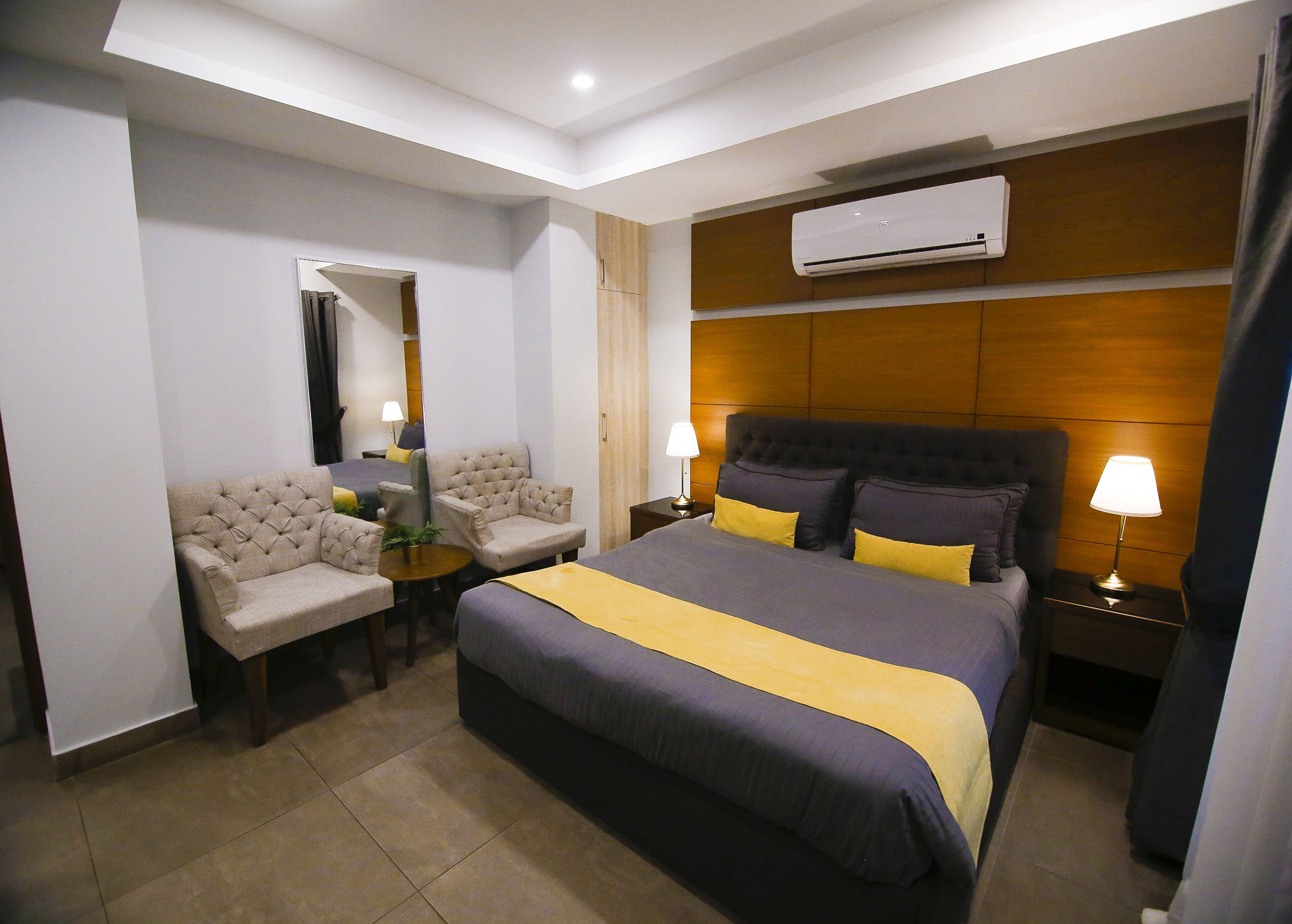 Habitación de hotel con cama, dos sillones, una mesita, unidad de aire acondicionado y mesita de noche con lámparas.