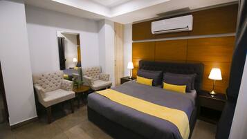 Habitación de hotel con cama, dos sillones, una mesita, unidad de aire acondicionado y mesita de noche con lámparas.