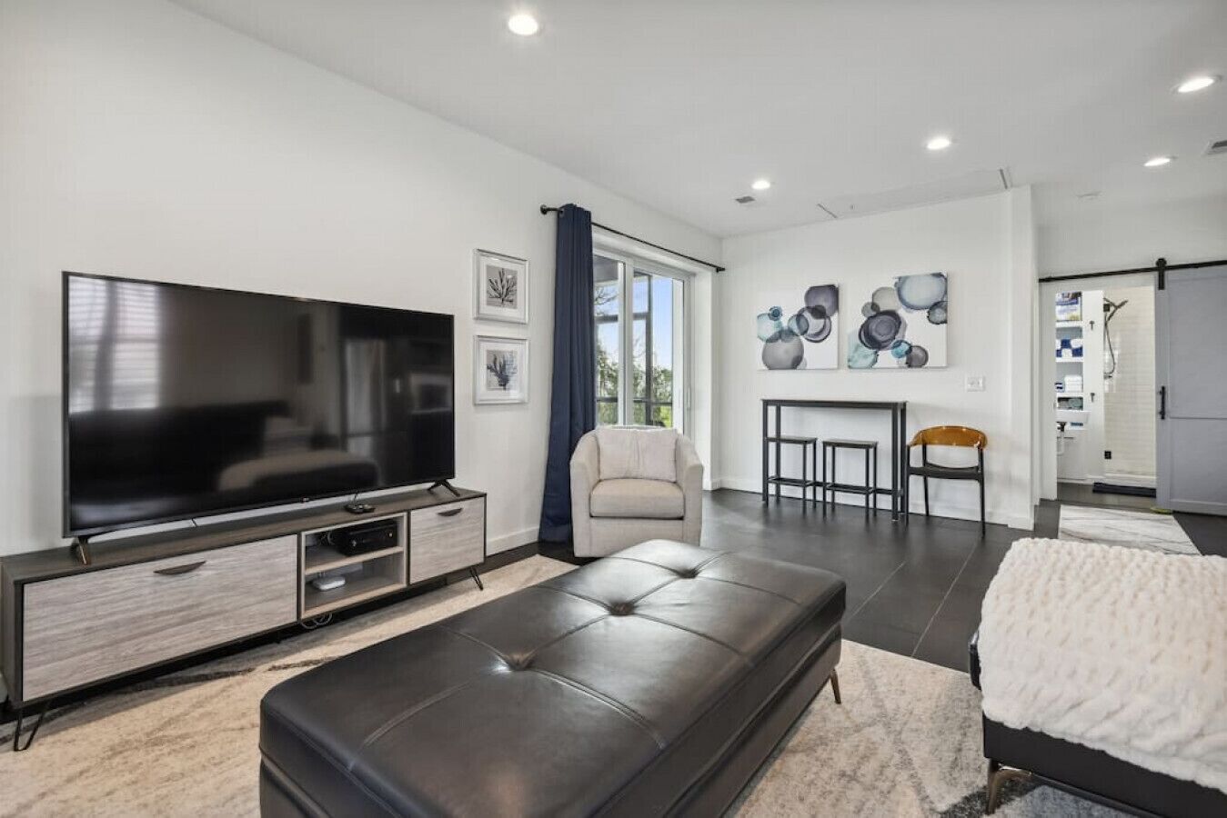 A modern living room with a large flat-screen TV, a leather sofa, a coffee table, and a dining area with a small table and chairs.