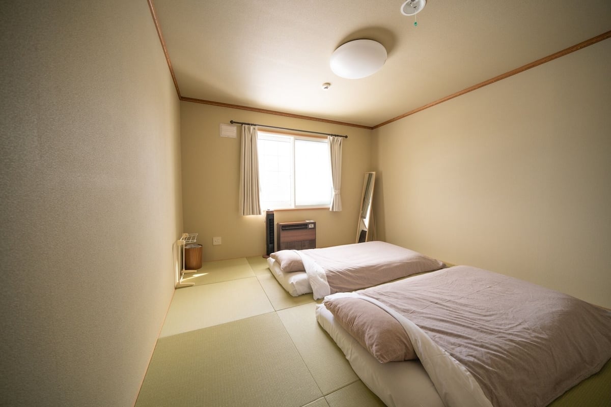A small, clean room with two beds, a window with curtains, a radiator, and a mirror on the wall.