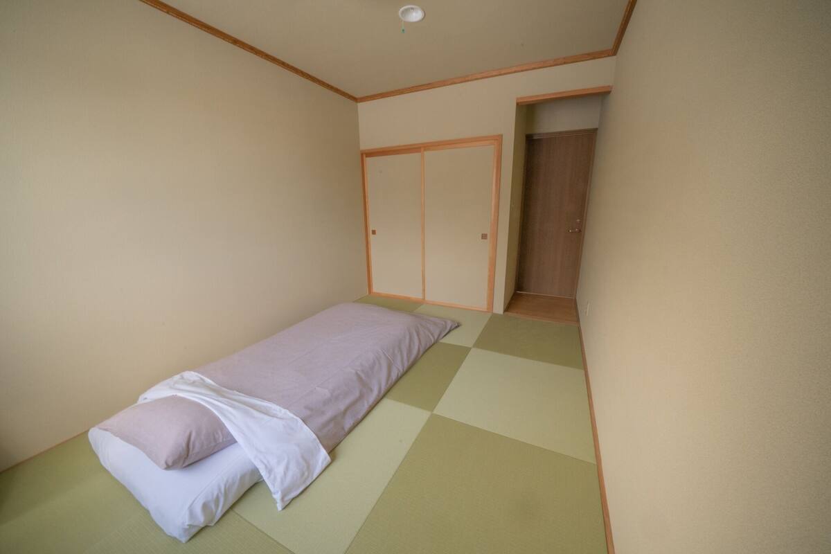 A small, single-bed room with a tatami mat floor, a wooden wardrobe, and a door leading to another room.