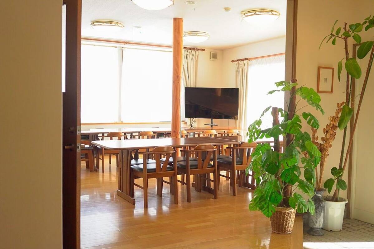 A dining area with wooden tables and chairs, a flat-screen TV, and large windows.