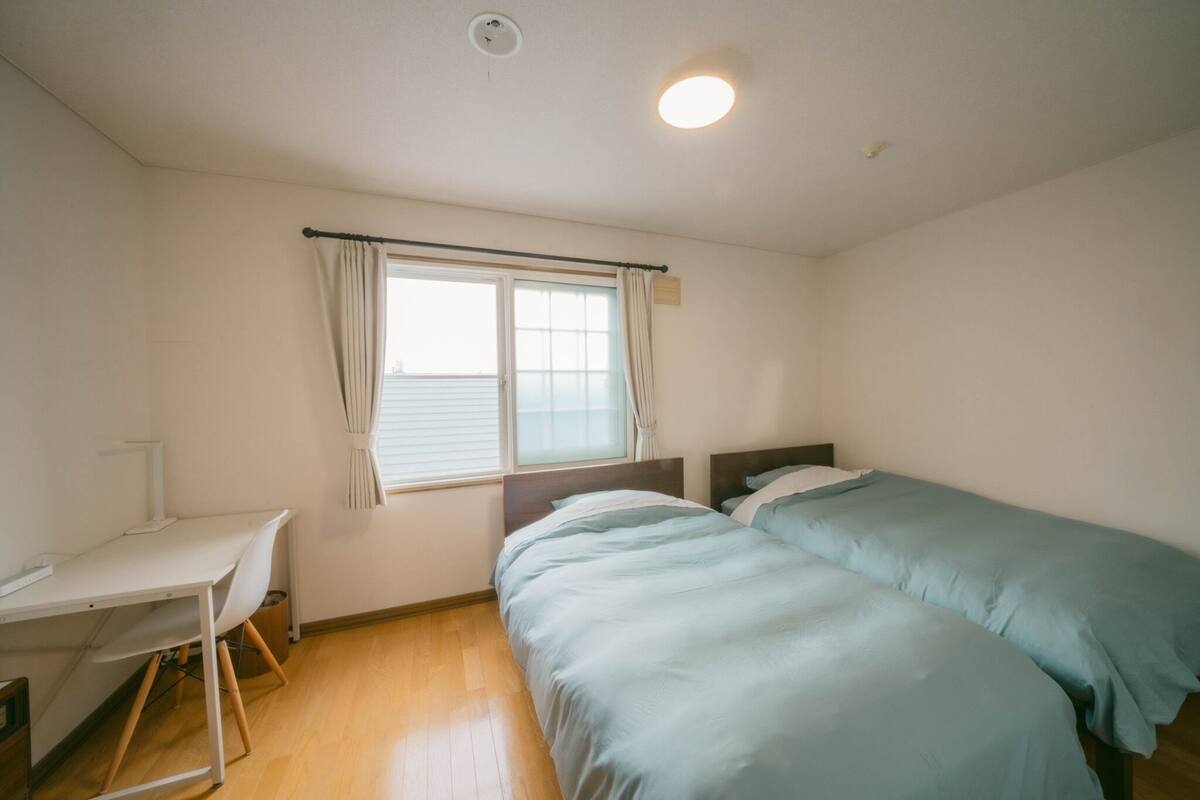 A bedroom with a desk, two beds, a window with curtains, and a ceiling light.
