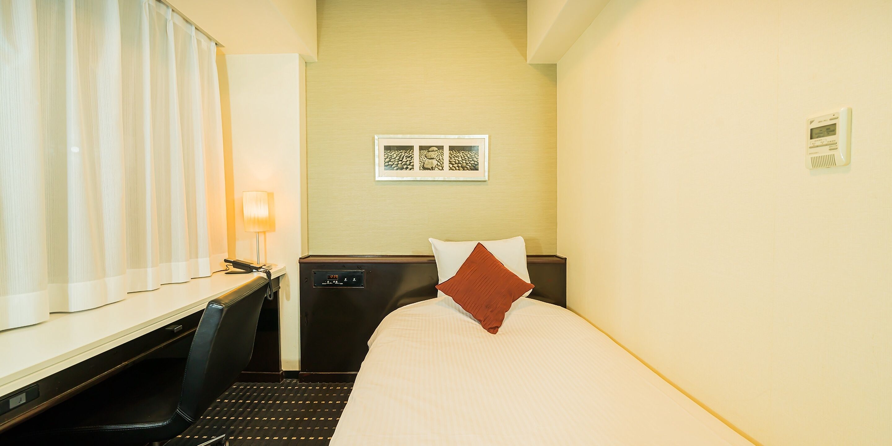 A hotel room with a bed, a desk, a chair, a lamp, and a thermostat on the wall.