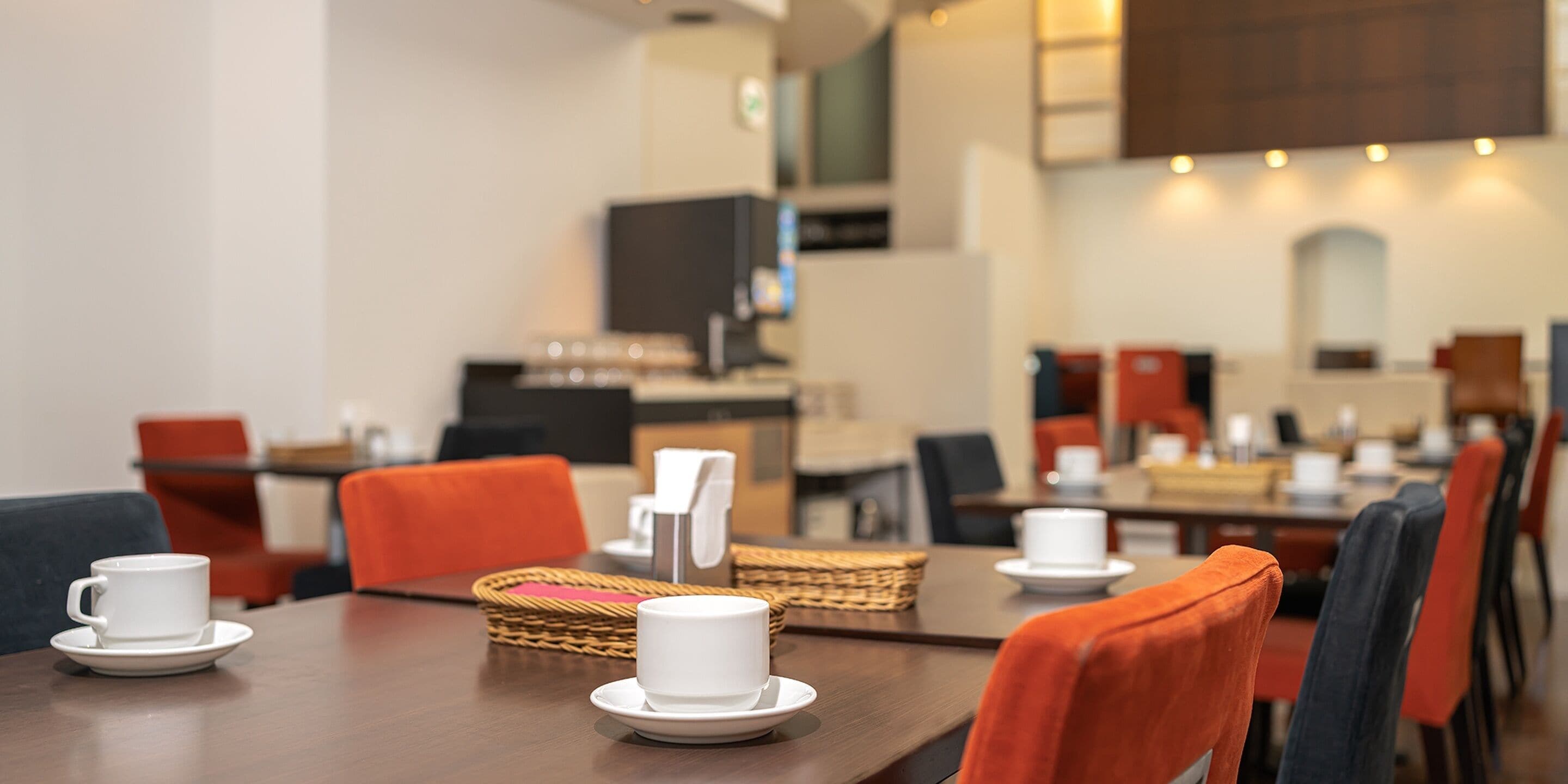 A dining area with tables set for two, each with a white cup and saucer, and orange chairs.