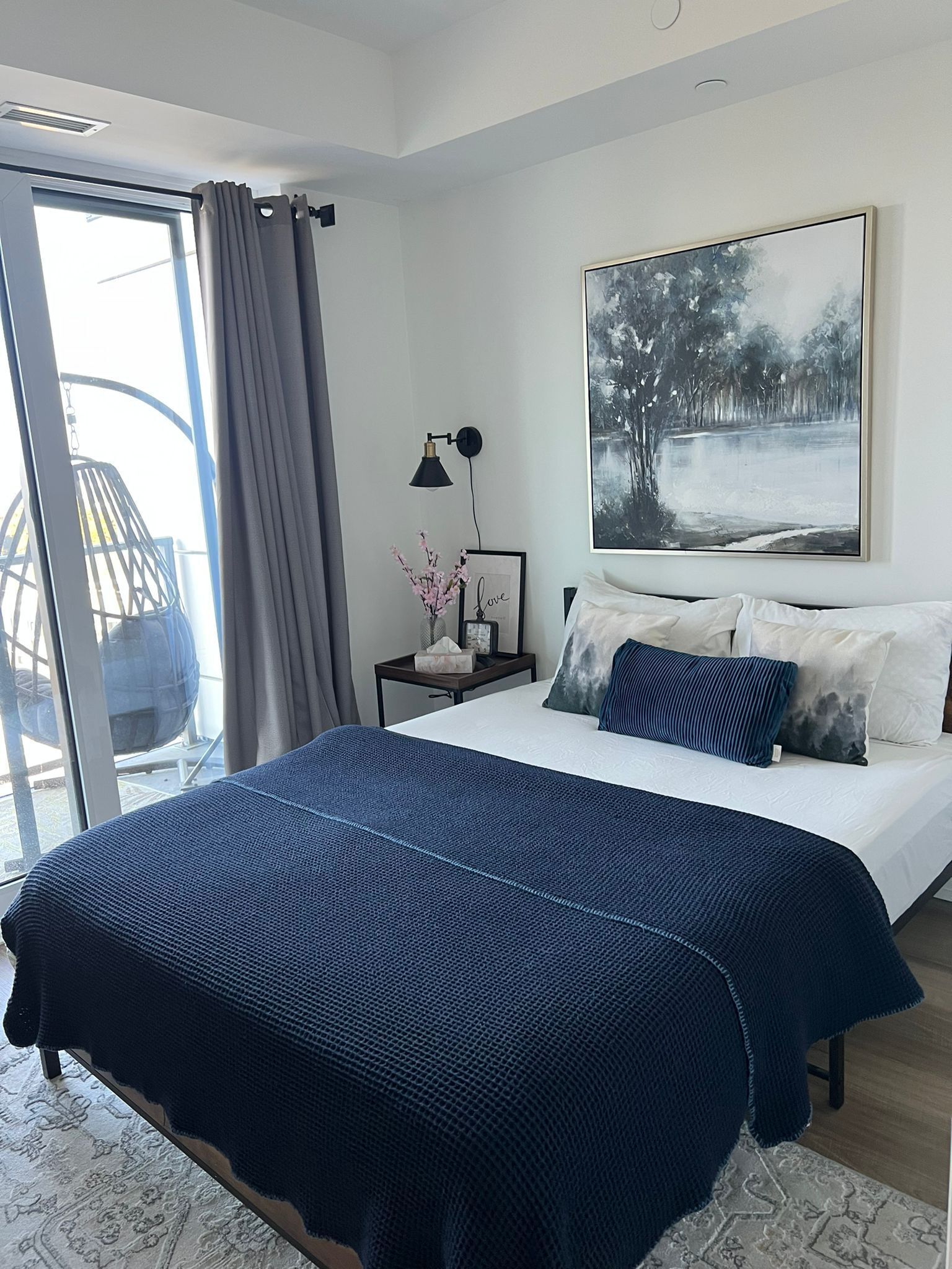 A modern bedroom with a large bed, a side table, a framed picture, and a view through a glass door.