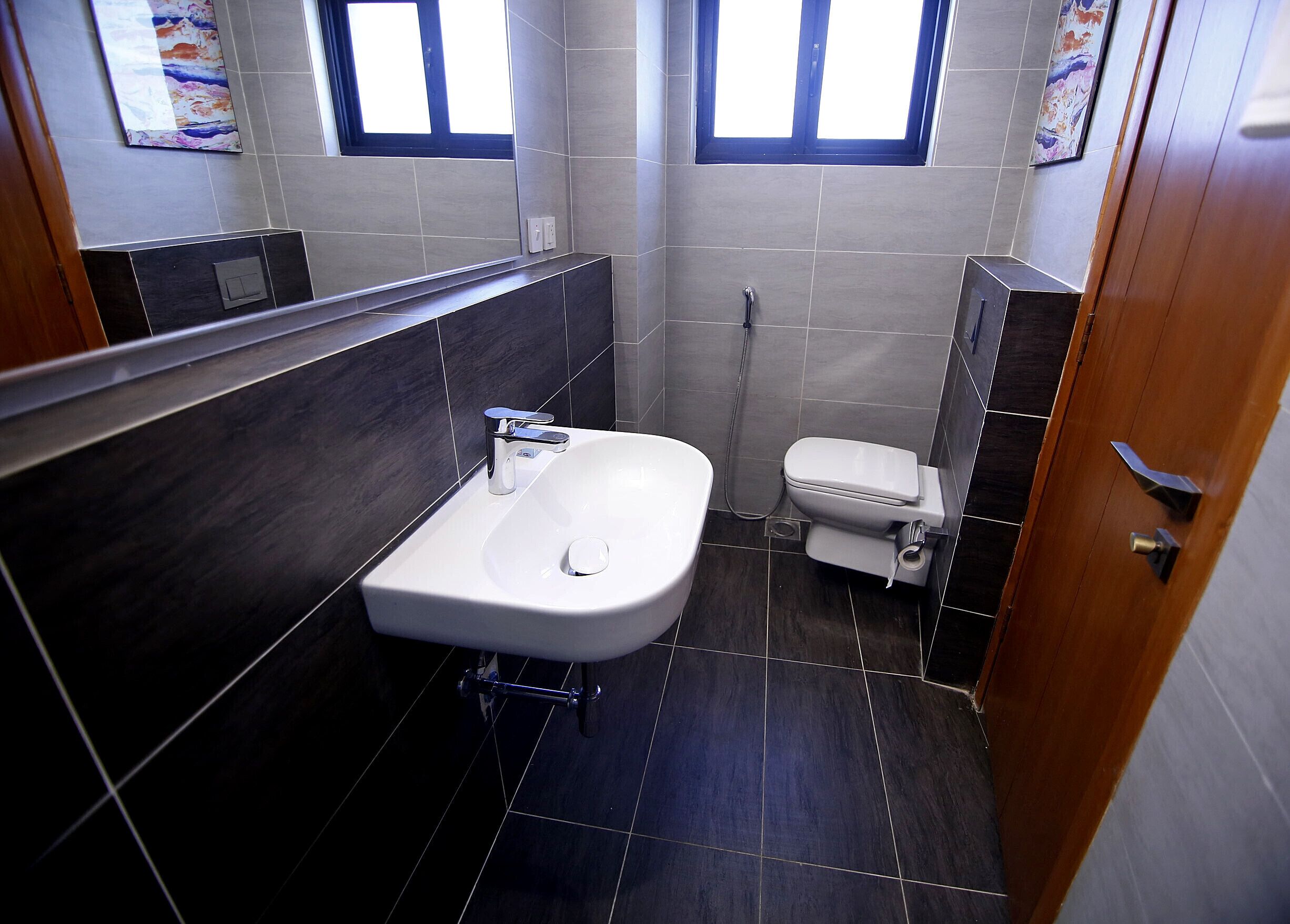 A bathroom with a white sink, a toilet, and a shower. There are two windows providing natural light.