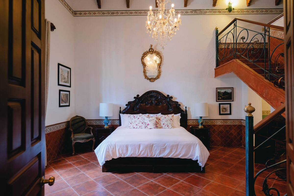 A bedroom with a large bed, a chandelier, a staircase, and a mirror on the wall.