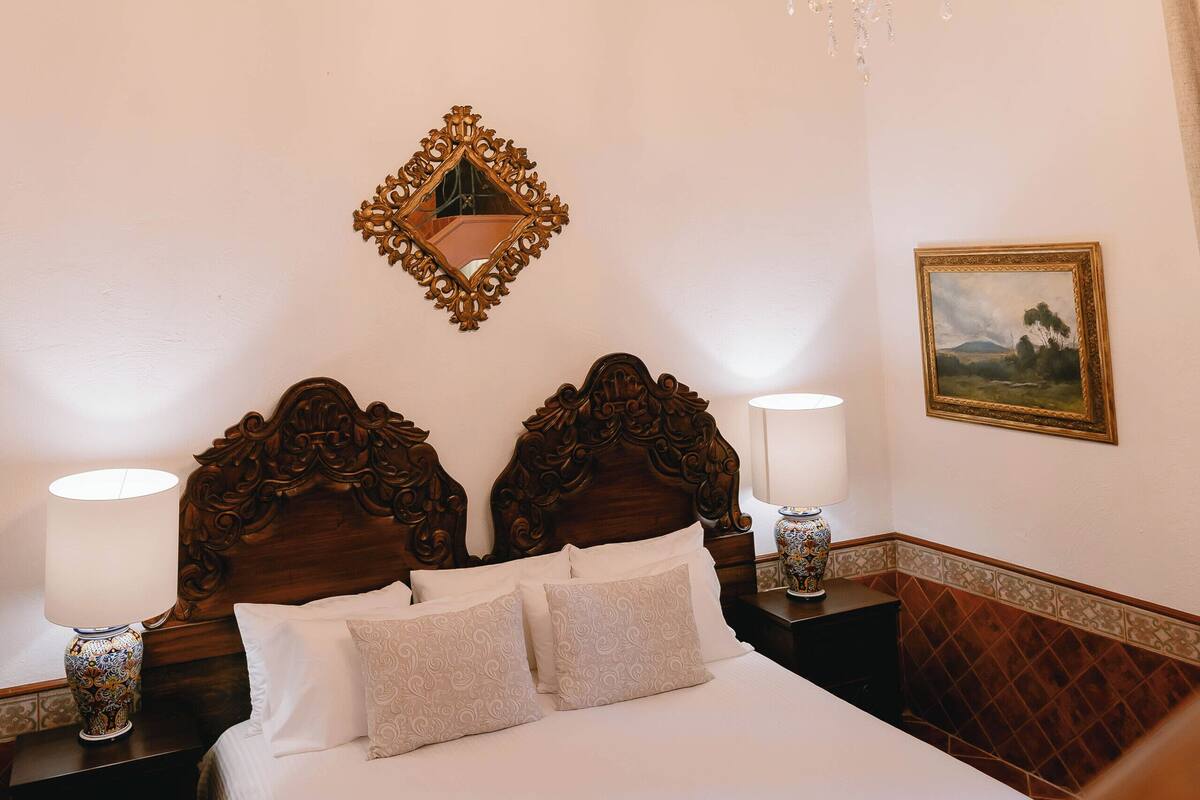 A bedroom with a wooden headboard, a chandelier, a bedside table with a lamp, and a framed picture on the wall.