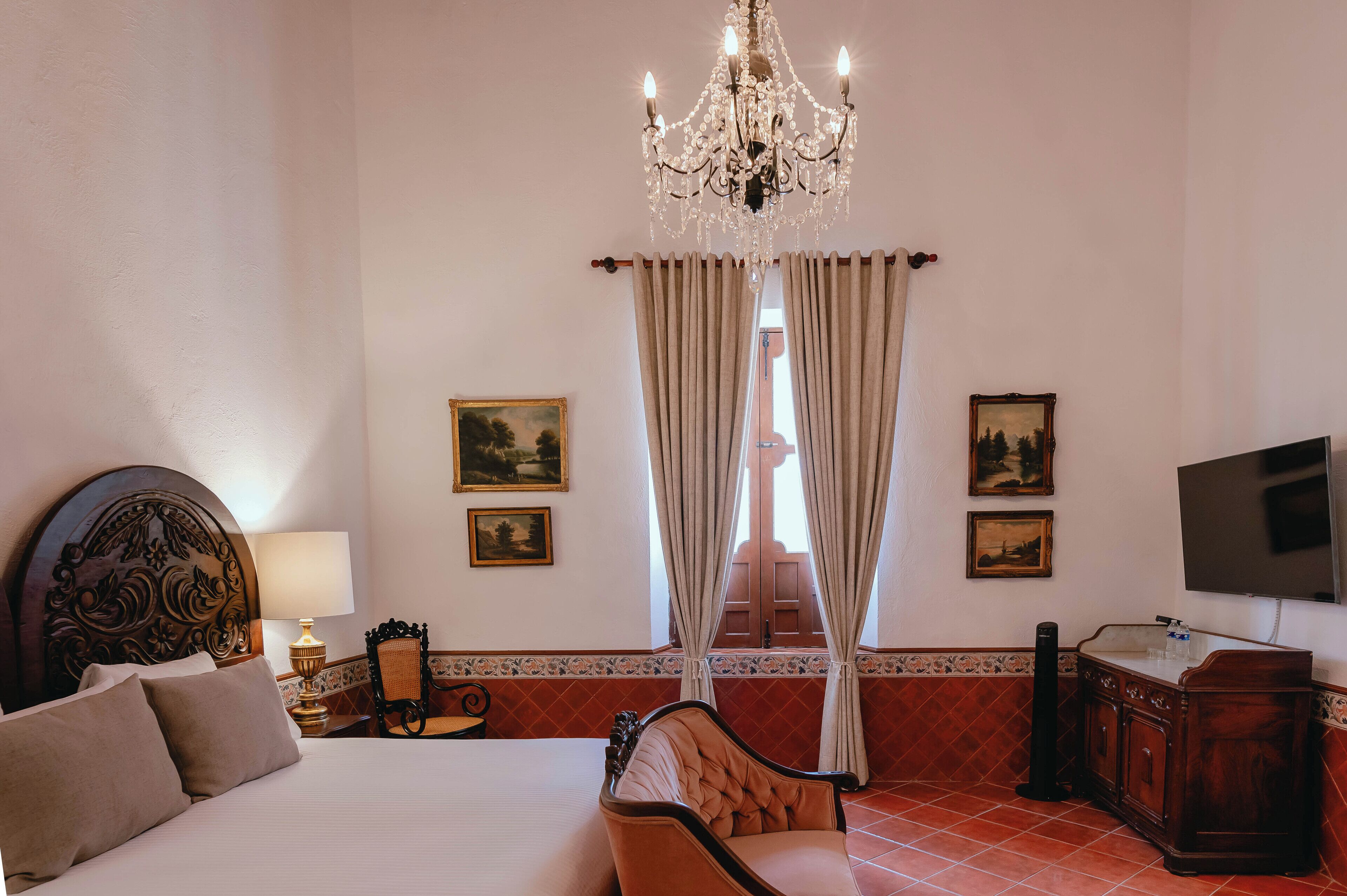 A hotel room with a bed, a chair, a desk, a TV, and a chandelier.