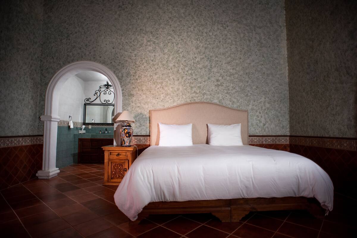 A bedroom with a large bed, a nightstand, a mirror, and a view of a bathroom.