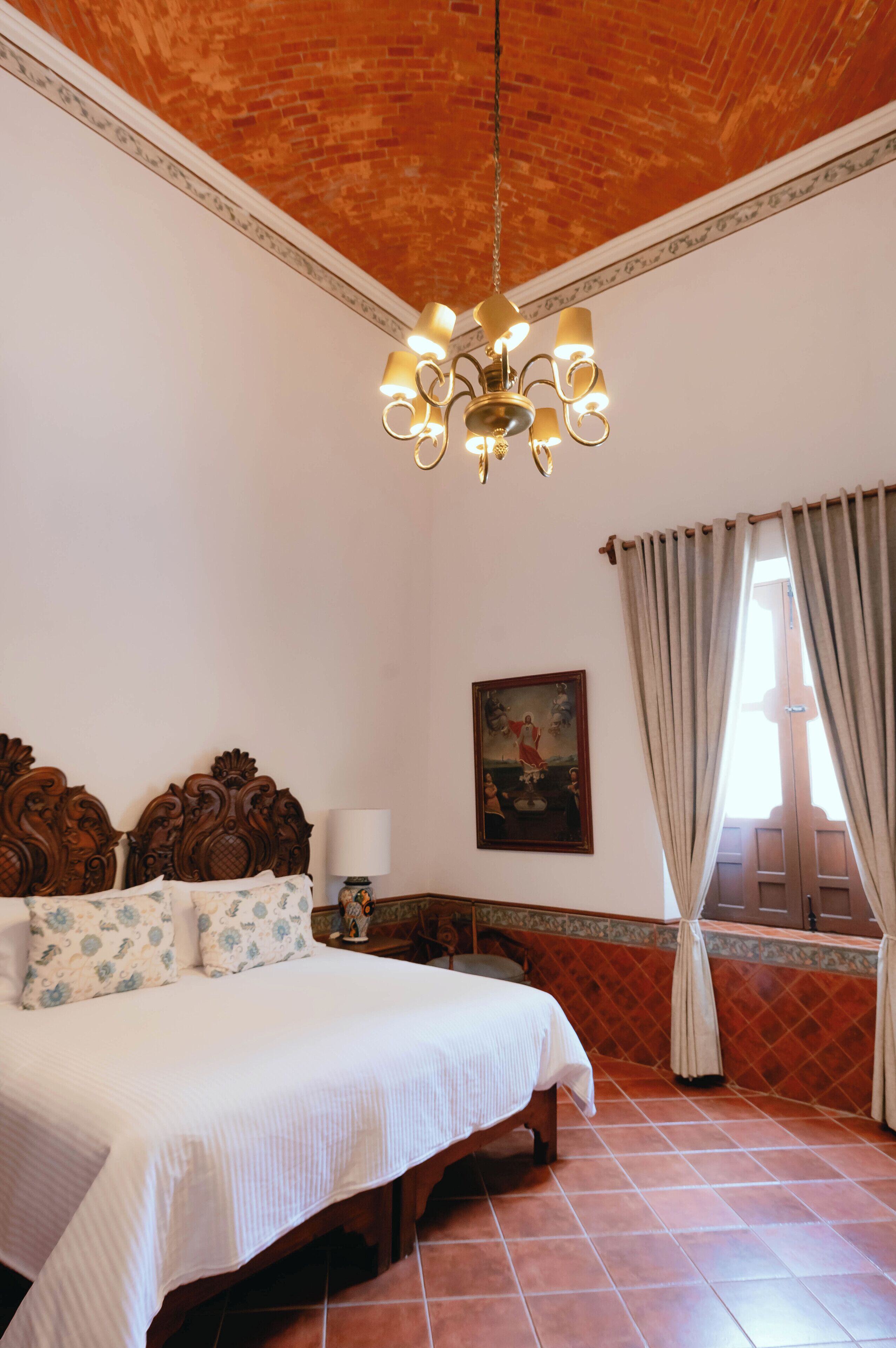 A bedroom with a large bed, a chandelier, a painting, and a window with curtains.