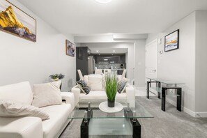Ett modernt vardagsrum med ett soffbord i glas, en vit soffa och ett kök i bakgrunden.