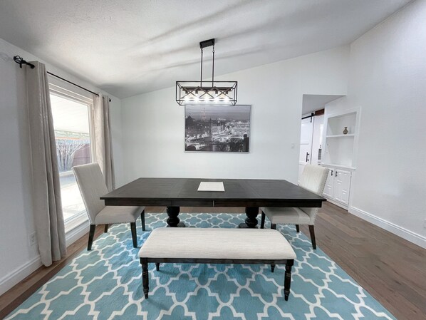 Un coin repas avec une table en bois foncé, deux chaises, un banc, un tapis à motifs, une œuvre d’art représentant un paysage urbain et un meuble blanc.