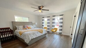 Une chambre à coucher avec un lit, un berceau en bois, un ventilateur de plafond et une fenêtre avec des rideaux.