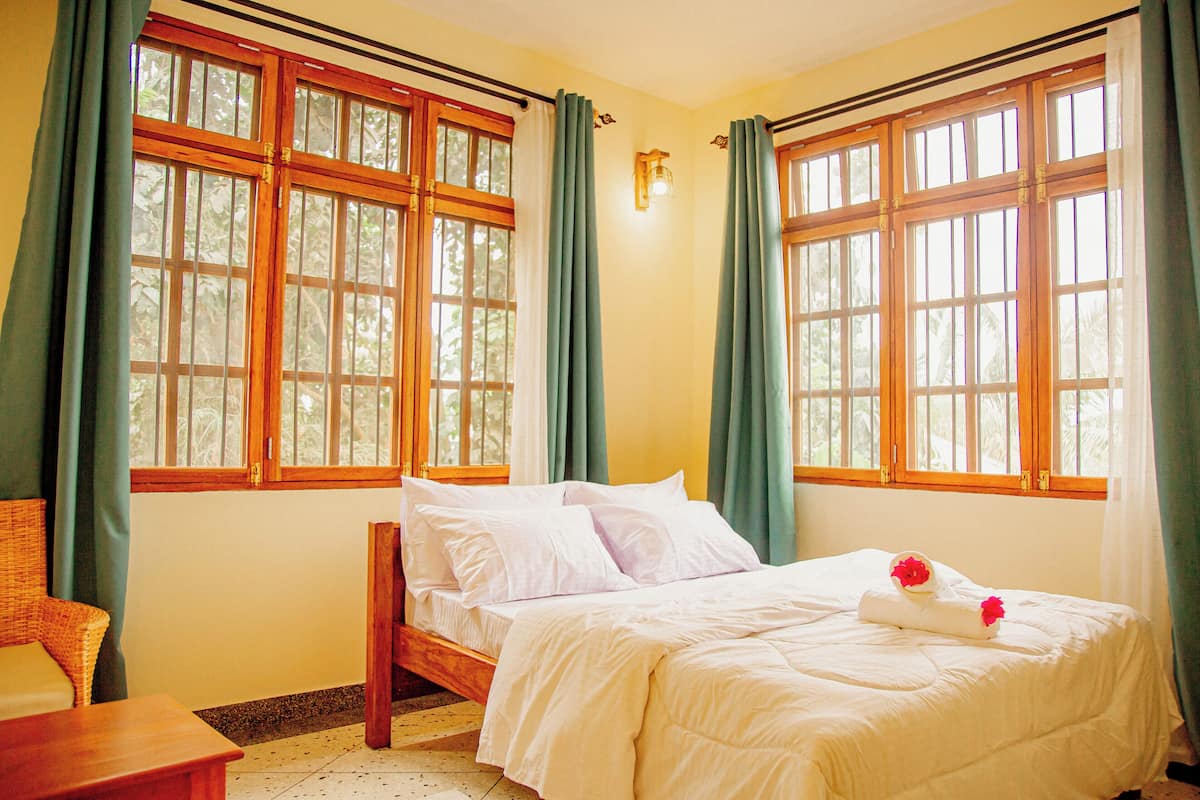 Un dormitorio con cama, una mesita de noche de madera, una silla y ventanas amplias con cortinas.